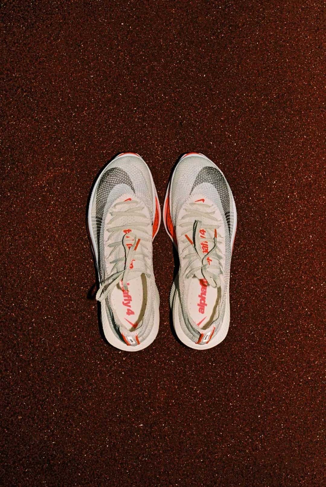 Par de Nike Alphafly 4 visto desde arriba sobre fondo de tierra rojiza, con capellada blanca tejida, cordones claros y el nombre del modelo en rojo en la plantilla