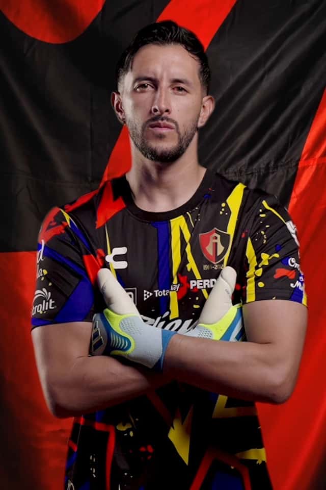 Camilo Vargas, arquero del Atlas, posa de brazos cruzados con un buzo de arquero de colores y guantes, delante de una bandera roja y negra; foto oficial de archivo de la temporada 2026 (foto: Atlas FC)