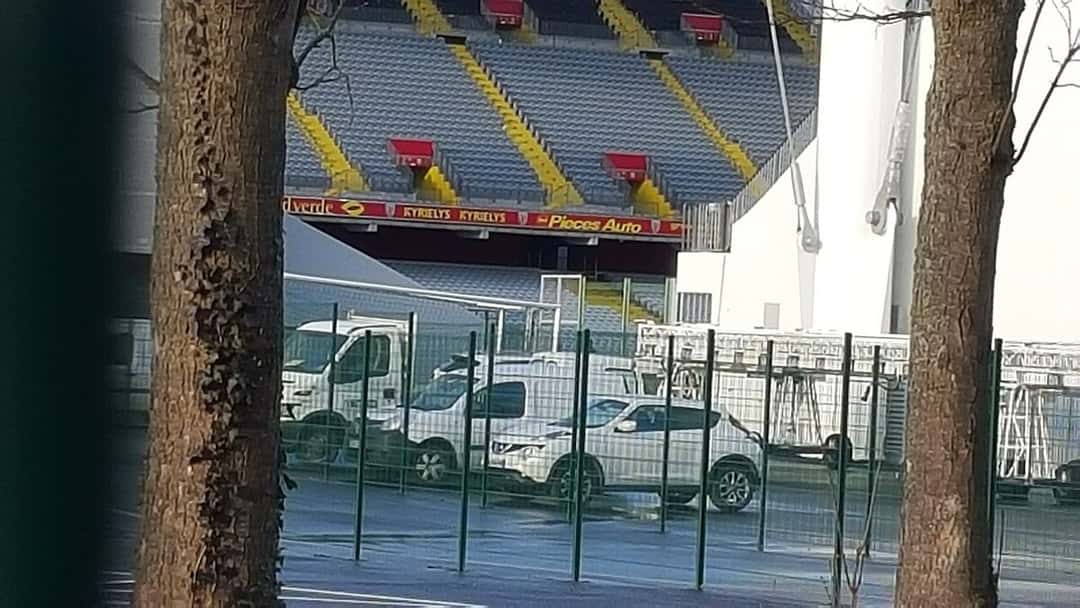 Una tribuna del estadio Bollaert-Delelis, con sus butacas amarillas y rojas, vista desde la calle a través de los árboles y el alambrado; foto de archivo de enero de 2016 (foto: Supporterhéninois, Wikimedia Commons, dominio público)