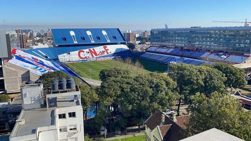 El Gran Parque Central visto desde la terraza de un edificio vecino, con la tribuna pintada con las letras del Club Nacional de Football y la cancha verde rodeada de árboles; foto de archivo de 2021 (foto: CotoBolso, Wikimedia Commons, licencia CC BY-SA 4.0)