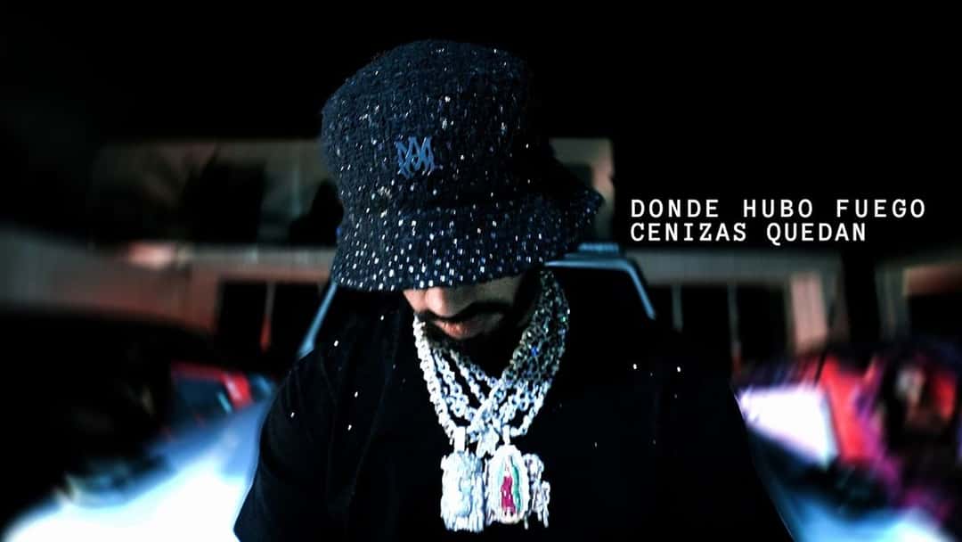 Fotograma de archivo del video oficial de «Donde Hubo Fuego Cenizas Quedan»: Anuel AA, con piluso negro brillante y cadenas con dijes de piedras, de frente a cámara, con el título de la canción en letras blancas