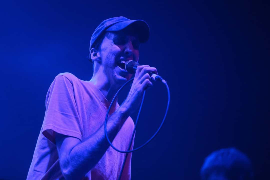 Bradford Cox canta con una gorra y una remera rosa en un show en Boston, iluminado en azul (foto de archivo, diciembre de 2015). Foto: Ben Stas, CC BY 2.0, vía Wikimedia Commons