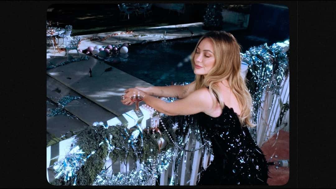 Fotograma del visualizer oficial de «I Guess it's Christmas»: Hilary Duff, con vestido negro brillante, apoyada en una baranda decorada con guirnaldas plateadas junto a una pileta