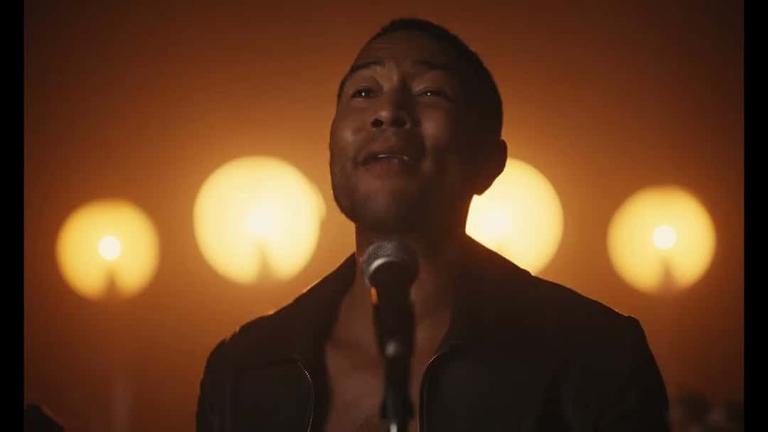 John Legend canta frente a un micrófono, con campera negra, delante de cuatro luces redondas anaranjadas, en un fotograma del video oficial de «Daylight»