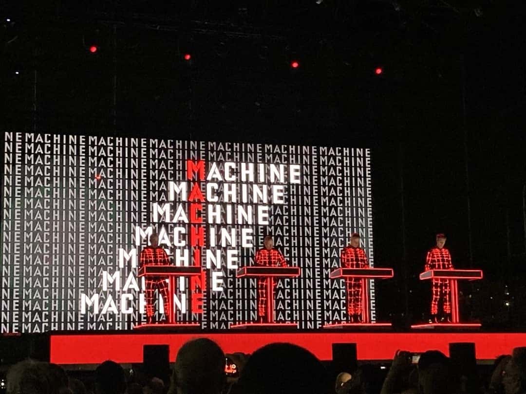 Kraftwerk en vivo en el Arena del Mare de Génova en julio de 2023: los cuatro integrantes con trajes rojos cuadriculados, detrás de sus consolas, frente a una pantalla llena de la palabra MACHINE en blanco y rojo (foto de archivo). Foto: Luca Dell'Orto, CC BY-SA 4.0, vía Wikimedia Commons