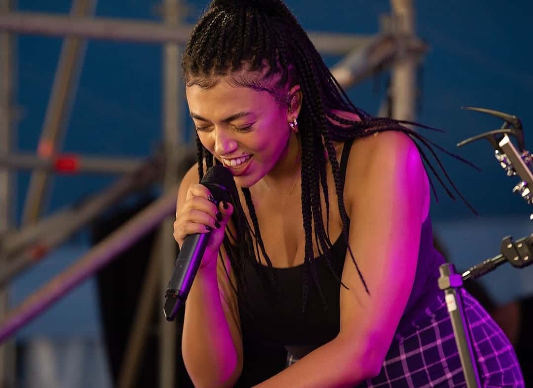 Mahalia canta inclinada hacia adelante, con trenzas largas y musculosa negra, sonriendo bajo luces violetas en un escenario al aire libre en Sídney (foto de archivo, enero de 2019). Foto: Bruce from Sydney, CC BY 2.0, vía Wikimedia Commons