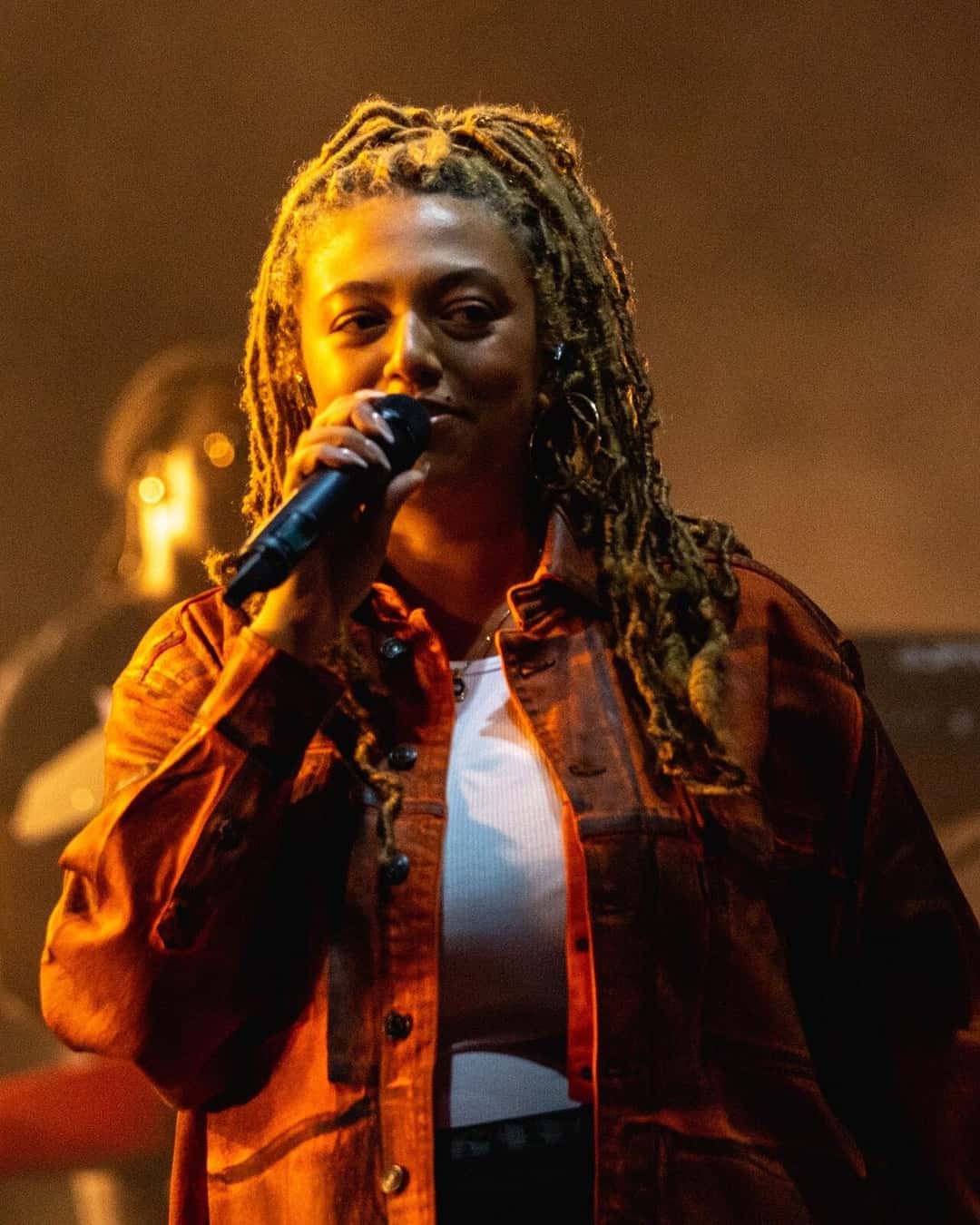 Mahalia canta con el micrófono cerca de la boca, con camisa naranja y musculosa blanca, bajo una luz cálida en el festival Boardmasters (foto de archivo, agosto de 2021). Foto: Raph_PH, CC BY 2.0, vía Wikimedia Commons