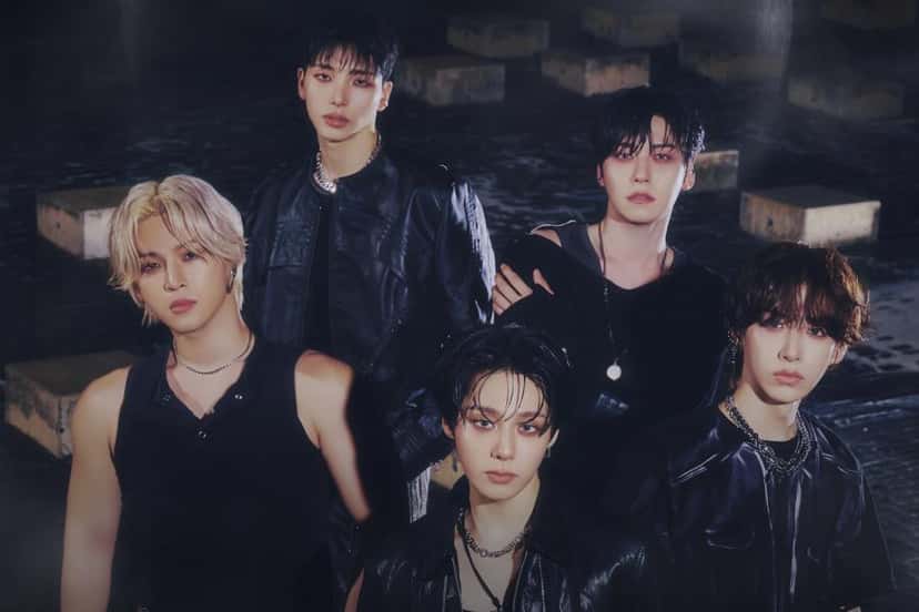 Los cinco integrantes de ONEUS con ropa negra de cuero, cadenas plateadas y maquillaje oscuro, sobre un piso mojado con bloques de cemento, en una foto oficial de B.WAVE Entertainment