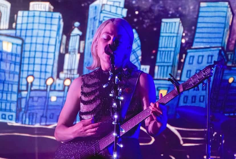 Phoebe Bridgers canta con los ojos cerrados y toca una guitarra brillante con luces en el pie del micrófono, frente a una pantalla con dibujos de edificios iluminados, en un show en Marymoor Park, Estados Unidos, en 2022 (foto de archivo). Foto: David Lee, CC BY 2.0, vía Wikimedia Commons