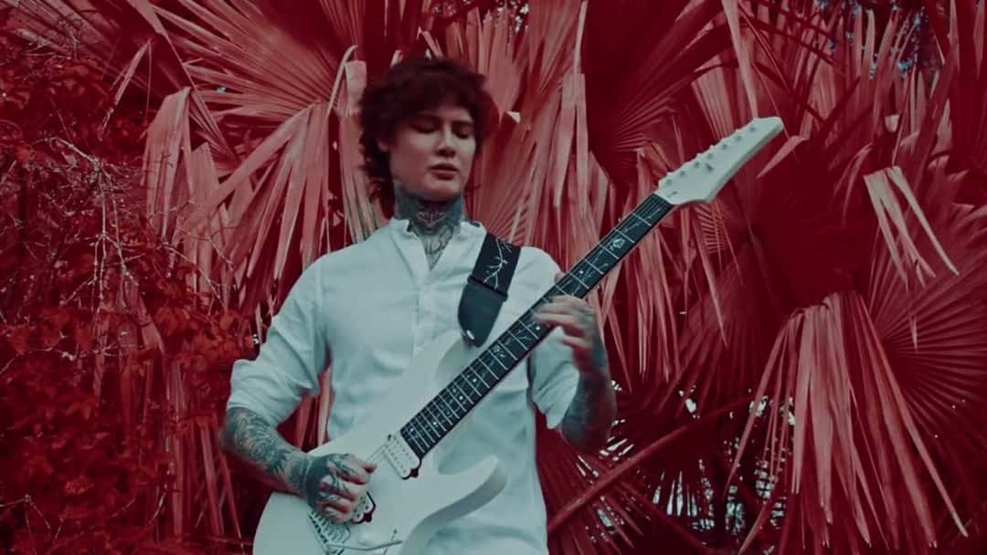 Un integrante de Polyphia, con camisa blanca y una guitarra eléctrica, rodeado de hojas de palmera rojas en el video oficial de «Power In The Blood» (fotograma del video oficial)