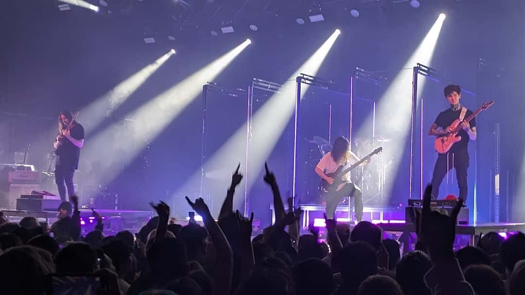 Polyphia en vivo en 2023 (foto de archivo): la banda toca bajo haces de luz blanca y violeta, con el público en primer plano. Foto: Makeworldpedia, CC BY-SA 4.0, vía Wikimedia Commons