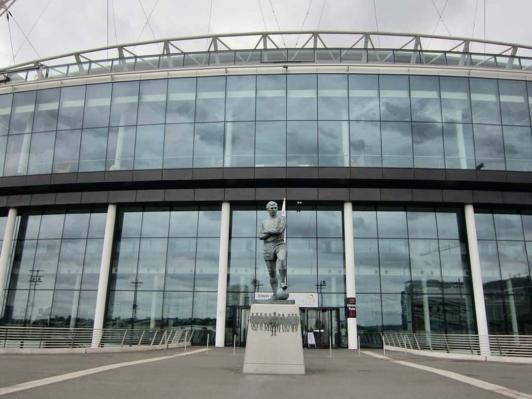 Fachada de vidrio del estadio de Wembley, en Londres, con la estatua de Bobby Moore frente a la entrada y el arco blanco arriba (foto de archivo). Foto: Ank Kumar, CC BY-SA 4.0, vía Wikimedia Commons