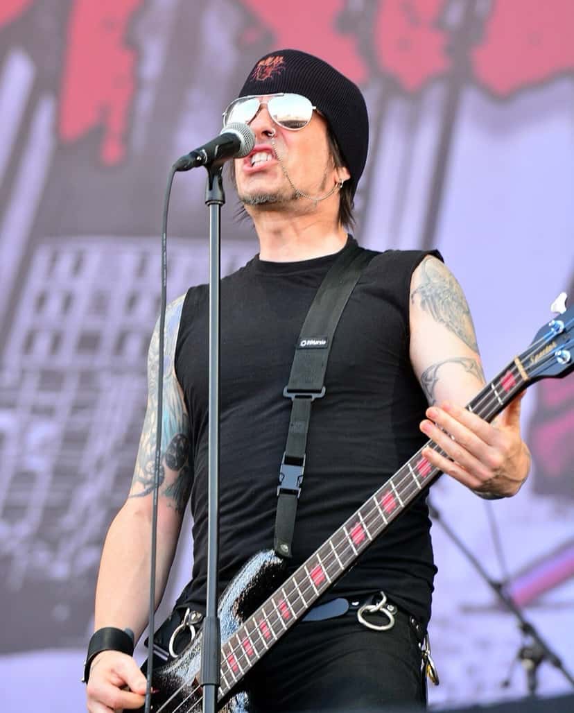 Rachel Bolan, bajista de Skid Row, canta en el micrófono mientras toca el bajo en el Wacken Open Air de 2014, con gorro negro y anteojos espejados (foto de archivo). Foto: P. Schwichtenberg, CC BY-SA 3.0, vía Wikimedia Commons