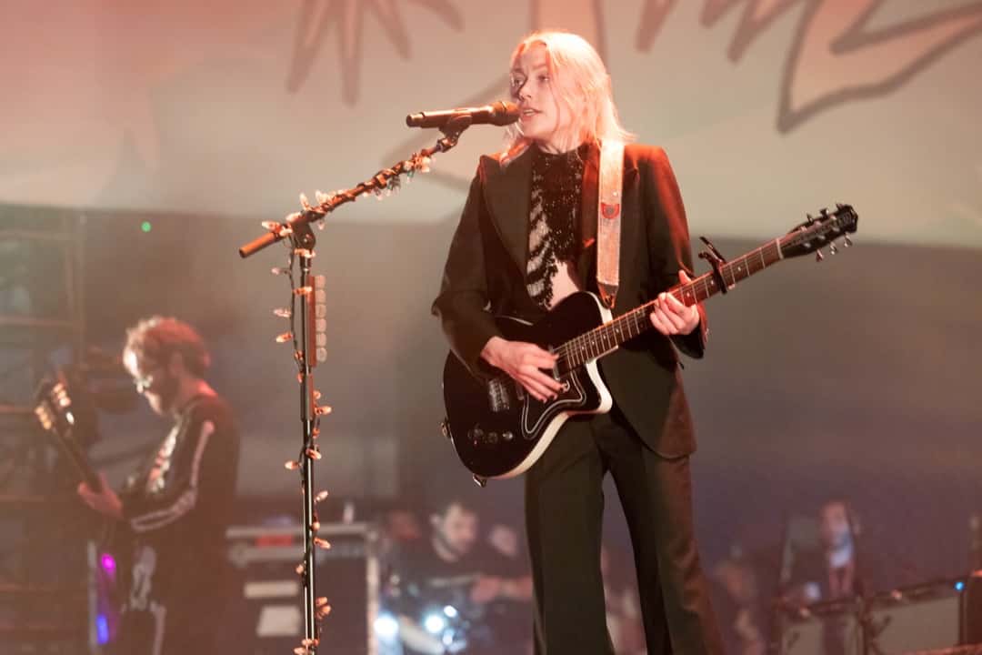 Phoebe Bridgers en vivo en el festival Laneway de Sídney en febrero de 2023: canta con pelo rubio platinado, traje oscuro y una guitarra negra, frente al micrófono (foto de archivo). Foto: Bruce Baker, CC BY 2.0, vía Wikimedia Commons