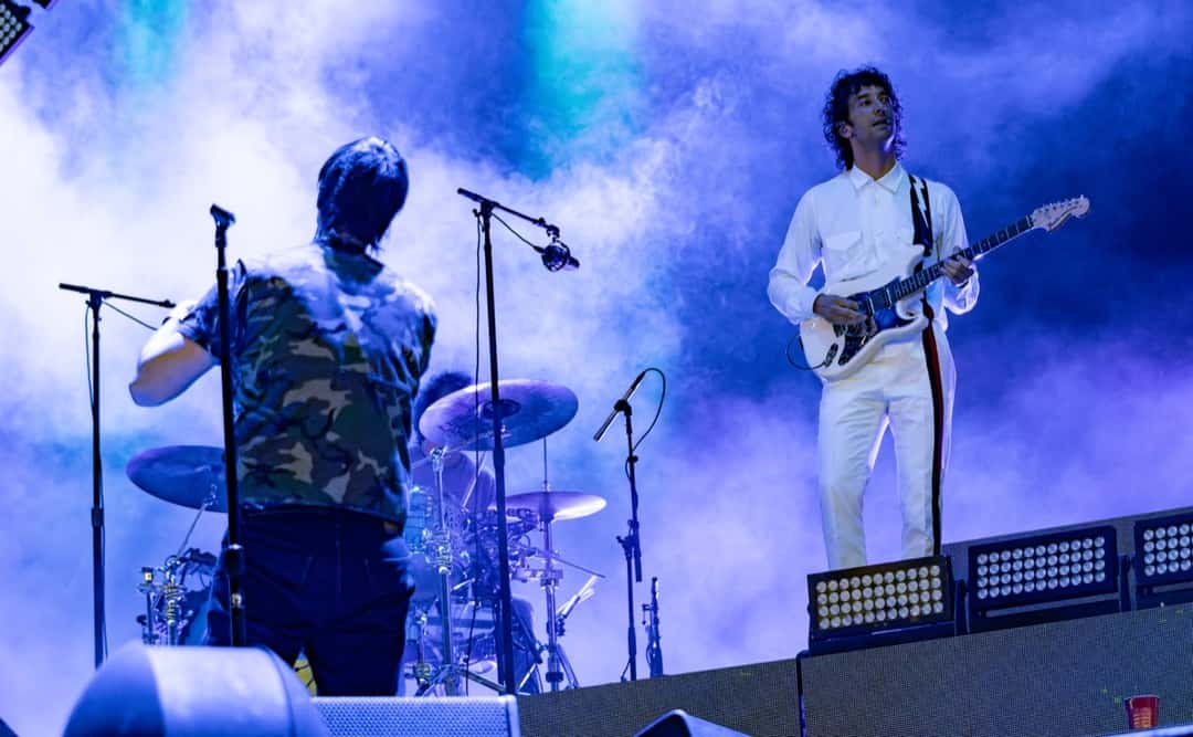 The Strokes en vivo en el NOS Alive de Lisboa en julio de 2022: Julian Casablancas, de espaldas y con camisa de camuflaje, frente a la batería, y un guitarrista vestido de blanco con una guitarra blanca, entre humo y luces azules (foto de archivo). Foto: Raph_PH, CC BY 2.0, vía Wikimedia Commons