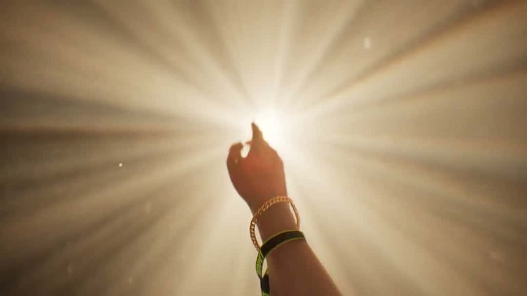 Fotograma del video oficial de «Chase The Feeling»: una mano con pulseras se estira hacia una luz blanca intensa que llena la pantalla