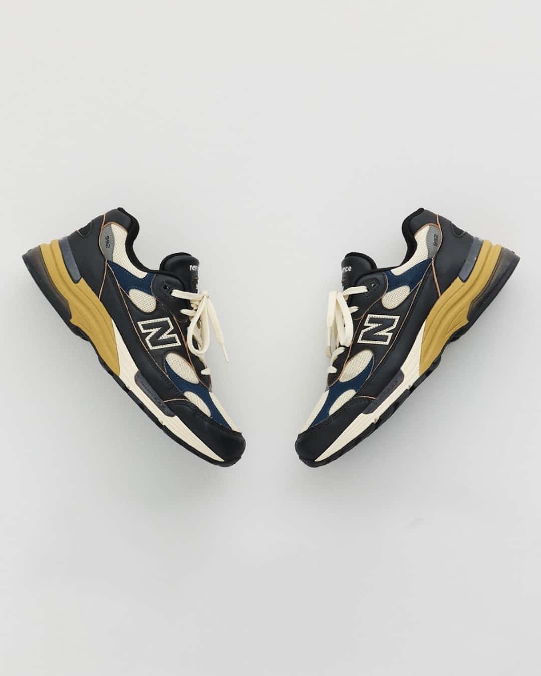 Par de New Balance 992 Made in USA Black/Afterglow enfrentados sobre fondo gris claro, con cuero negro, apliques azul marino, malla crema y el talón de la mediasuela en tono mostaza