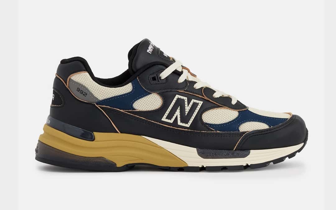 New Balance 992 Made in USA Black/Afterglow de perfil sobre fondo gris claro, con cuero negro, malla crema, apliques azul marino, la N en crema y la mediasuela crema con el talón en tono mostaza