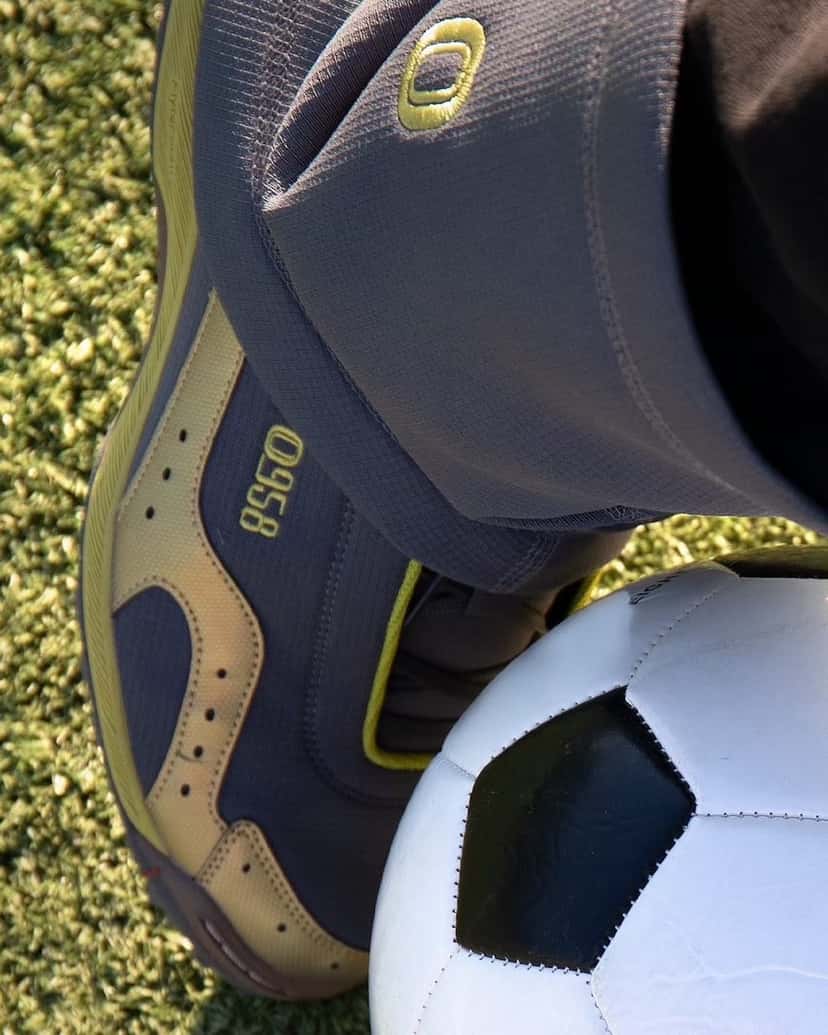 Detalle de la ASICS GEL-BELARRAOTTO de OTTO 958 con el pie apoyado sobre una pelota de fútbol, con la capellada gris oscura, el logo OTTO 958 en el lateral, detalles dorados y la polaina gris encima