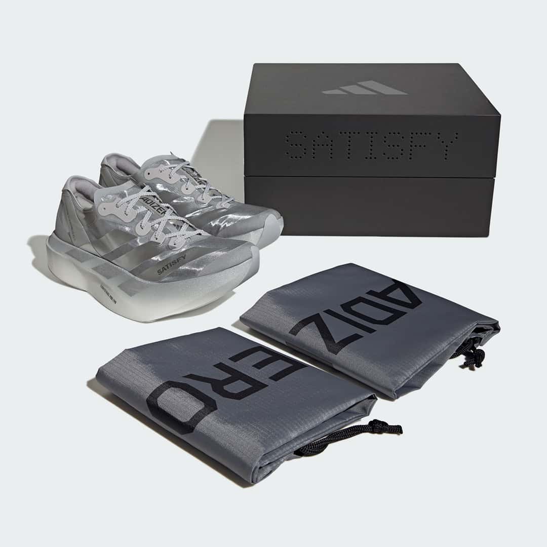Satisfy x adidas Adios Pro Evo 2 plateadas junto a su caja negra con la palabra SATISFY y dos bolsas grises con la inscripción ADIZERO