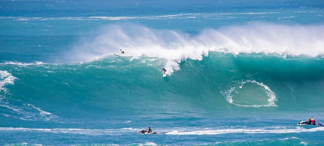 Un surfista baja una pared de agua gigante en Waimea Bay, con la ola rompiendo detrás y una moto de agua en el canal, en The Eddie de 2016 (foto de archivo)