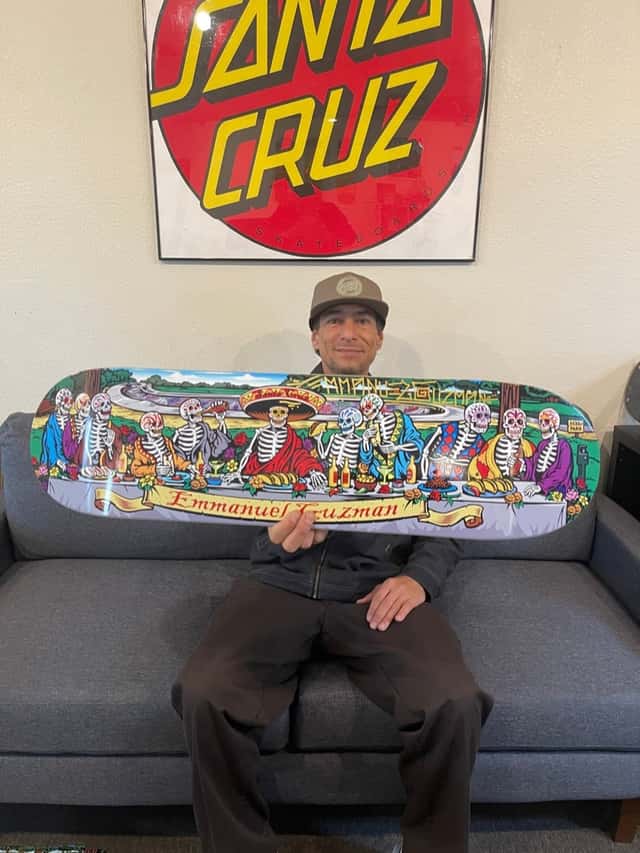 Emmanuel Guzmán, con gorra marrón, sentado en un sillón gris sostiene su tabla «Derby with the Dead», ilustrada con calaveras sentadas a una mesa larga, debajo de un cuadro con el logo de Santa Cruz