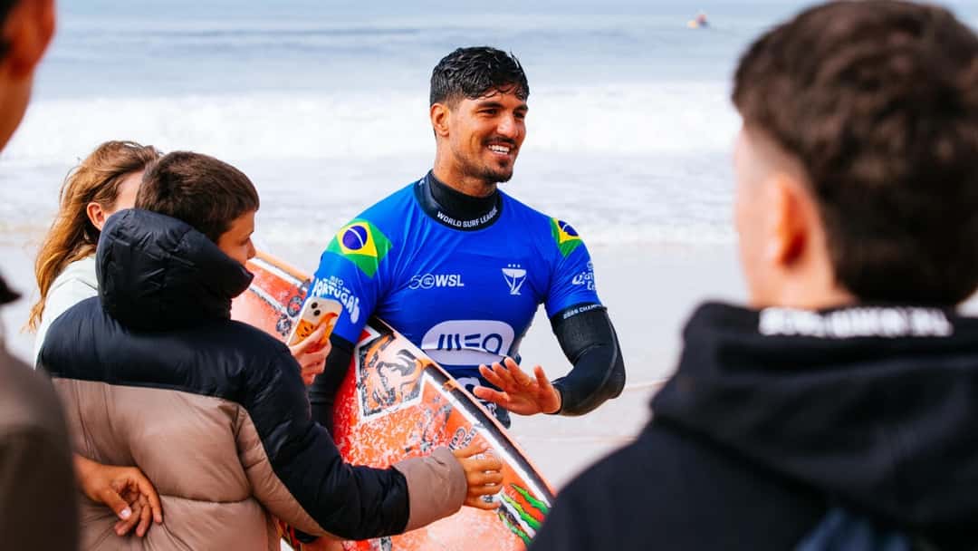 Gabriel Medina, con la lycra azul del MEO Rip Curl Pro Portugal y la bandera de Brasil, sonríe con su tabla mientras lo saludan en la orilla de Supertubos