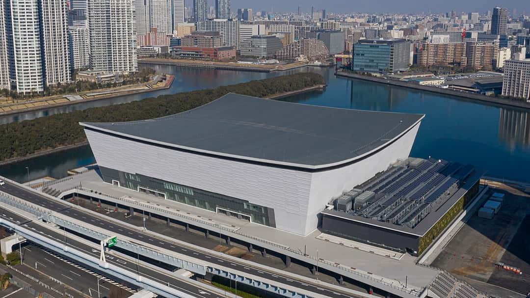 El Ariake Arena visto desde el aire, con su techo curvo gris, junto a un canal y rodeado de edificios de la bahía de Tokio