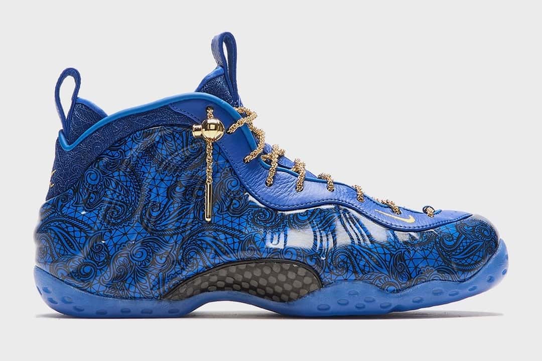 Victor Solomon x Nike Air Foamposite One Blue Trophies sobre fondo gris claro, con la carcasa azul con dibujo ornamental, cordones de soga dorados y el dije colgante
