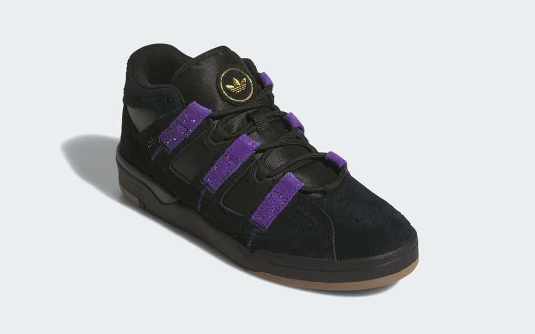 adidas Kader negro visto de frente en diagonal sobre fondo gris claro, con lengüeta de nylon negro y un parche redondo con el Trefoil dorado, cordones negros que pasan por las tiras de gamuza violeta moteada, puntera perforada y suela de goma color caramelo