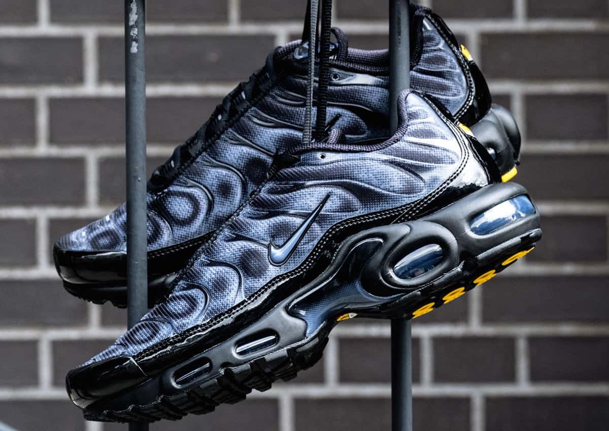Par de Nike Air Max Plus negro y gris colgado de una baranda frente a una pared de ladrillos, con el upper en degradé de puntos, las ondas laterales, la burbuja de aire azulada y detalles amarillos en la suela