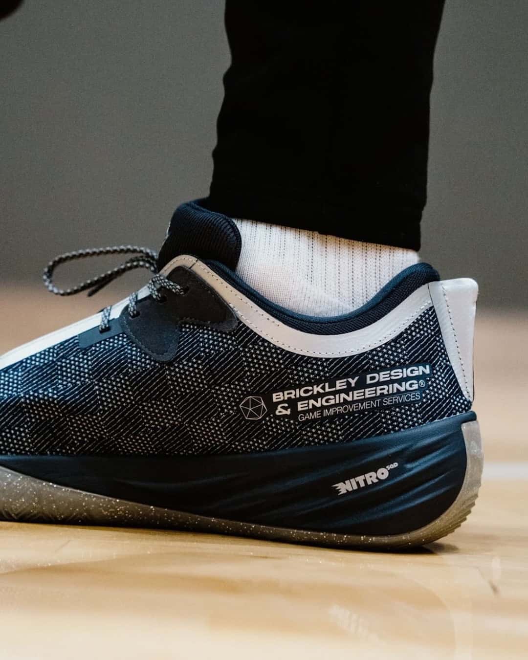 Costado de la PUMA All-Pro Nitro 2 Race 2 Evolve de Chris Brickley puesta en una cancha de madera, con la leyenda Brickley Design & Engineering, el logo Nitro en el talón y la suela gris con puntitos