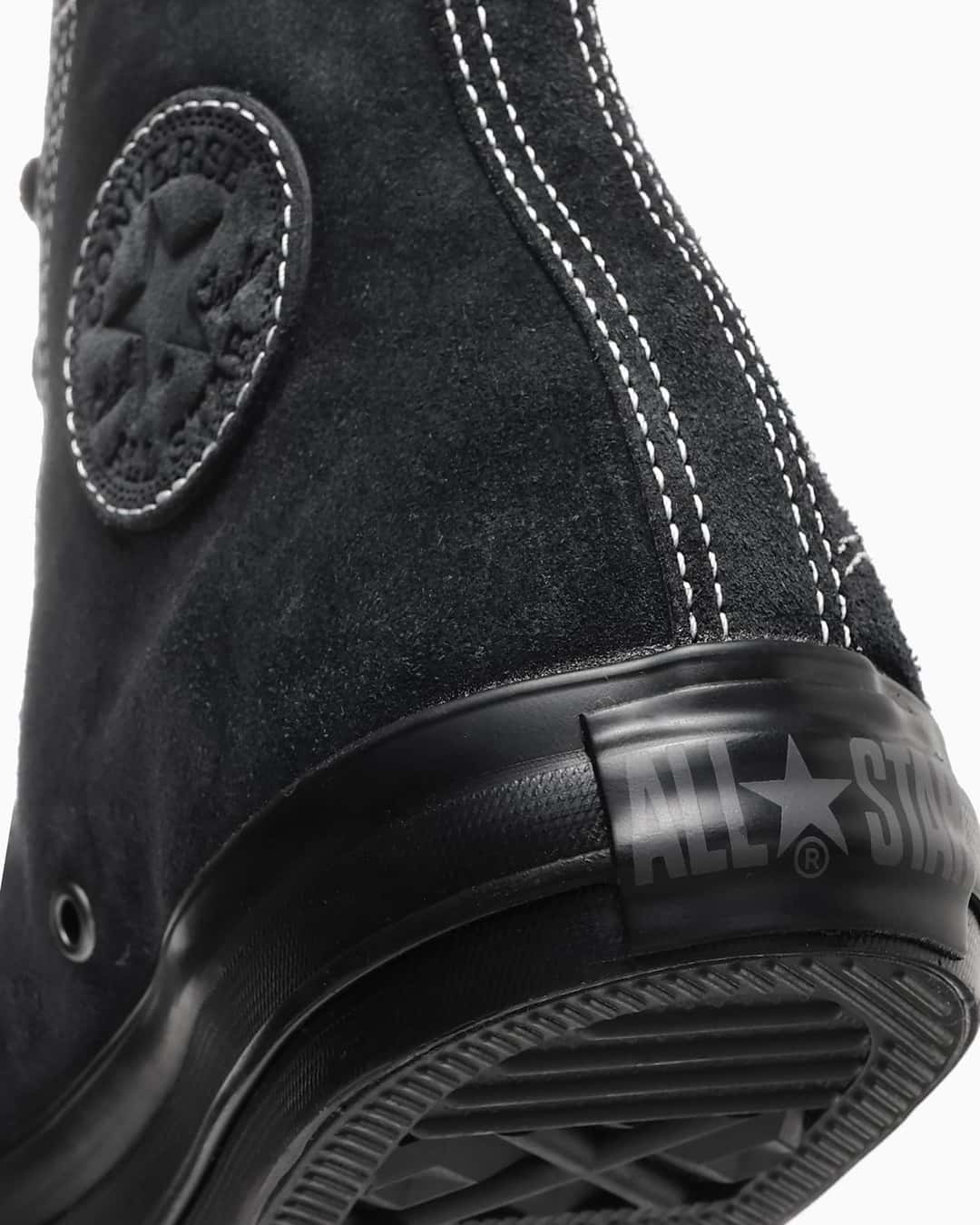 Primer plano del talón de la versión negra: gamuza negra con pespuntes blancos, el parche circular del tobillo en relieve y la etiqueta de goma del talón con el logo ALL STAR en gris sobre la suela negra