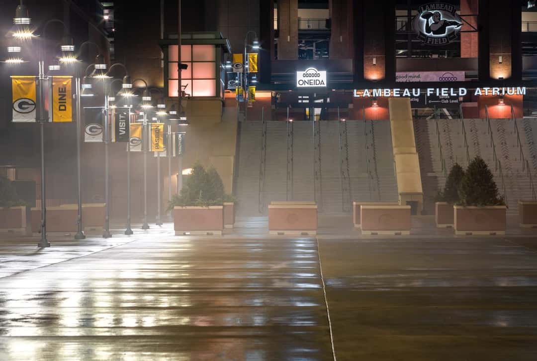 Imagen del estadio: la entrada del atrio del Lambeau Field de noche, con la calle mojada y los carteles de los Packers iluminados