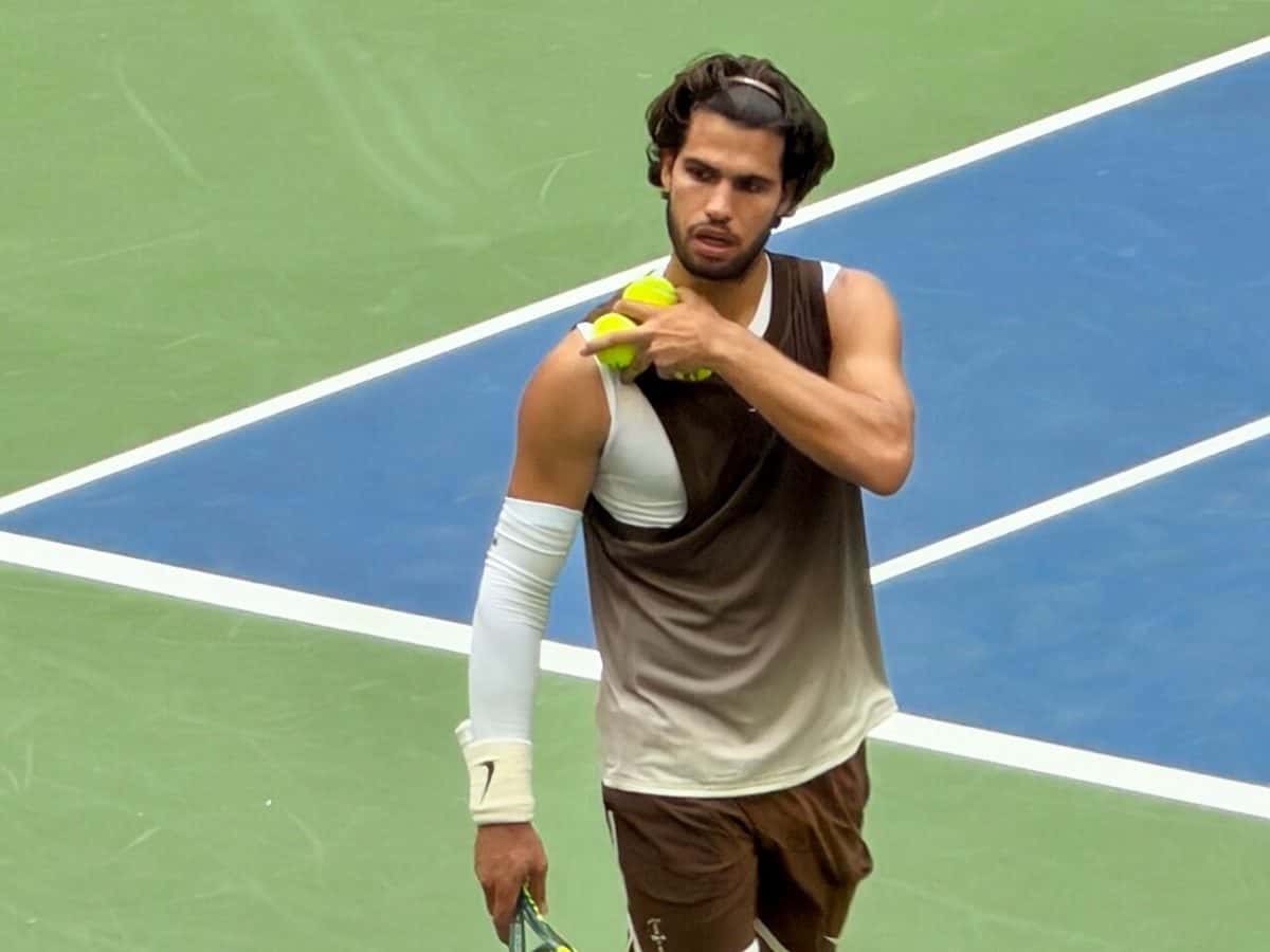 Foto de archivo: Carlos Alcaraz, con musculosa marrón y blanca, camina por una cancha dura azul y verde con pelotas en la mano en el US Open 2026