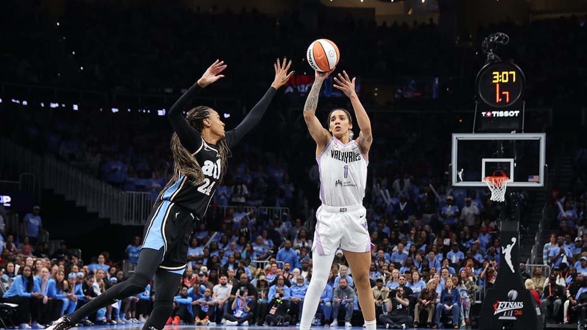 Gabby Williams, de las Valkyries, con la camiseta blanca número 1, lanza al aro ante una jugadora del Dream, de negro, con la tribuna celeste del State Farm Arena de fondo, en el Juego 3 de las Finales