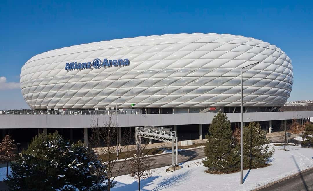 El Allianz Arena de Múnich, el estadio del Bayern, con su fachada blanca de paneles inflables, en un día de sol con nieve alrededor; foto de archivo de febrero de 2013 (foto: Diego Delso, Wikimedia Commons, licencia CC BY-SA 3.0)