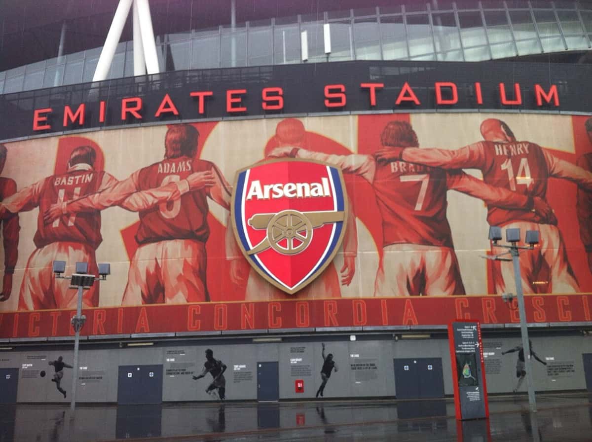 El escudo del Arsenal y el mural de leyendas del club en la fachada del Emirates Stadium, con el cartel del estadio arriba; foto de archivo de abril de 2016 (foto: Photo2023, Wikimedia Commons, dominio público, CC0)
