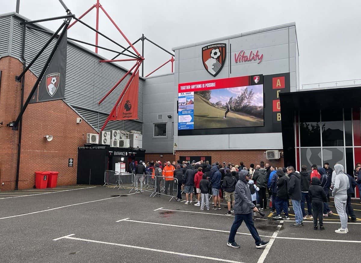 La entrada del Vitality Stadium de Bournemouth, con el escudo del club en la fachada, una pantalla y una fila de hinchas en la vereda; foto de archivo de 2023 (foto: Mr Ignavy, geograph.org.uk, Wikimedia Commons, licencia CC BY-SA 2.0)