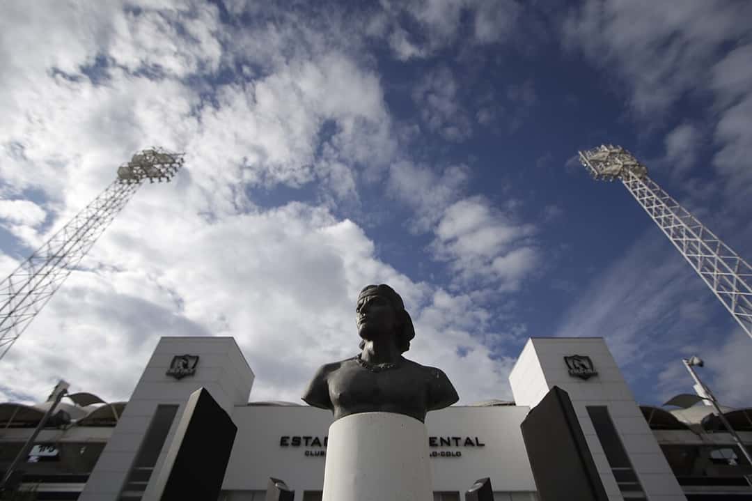 El busto de David Arellano frente a la entrada del estadio Monumental, con las torres de iluminación y el cielo con nubes de fondo; foto de archivo de junio de 2016 (foto: Ministerio Secretaría General de Gobierno, Wikimedia Commons, licencia CC BY-SA 2.0)