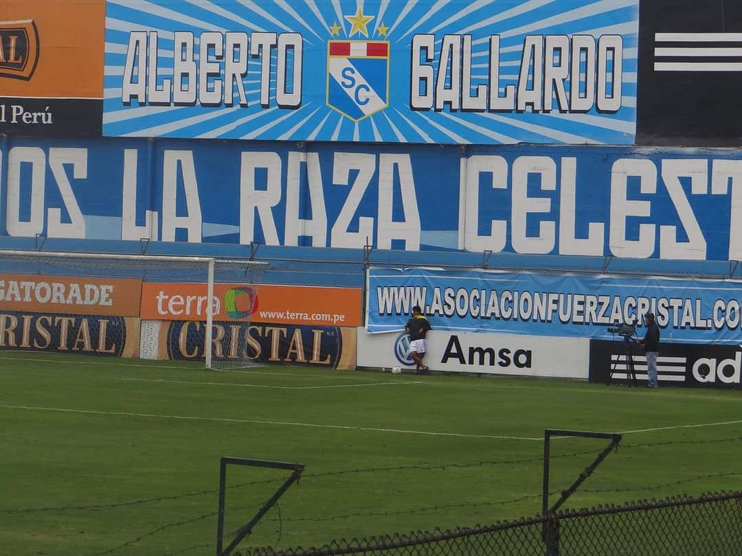 El cartel del estadio Alberto Gallardo, con el escudo de Sporting Cristal sobre un fondo celeste, y una tribuna con la leyenda de la hinchada; foto de archivo de julio de 2013 (foto: SCellar, Wikimedia Commons, licencia CC BY-SA 3.0)