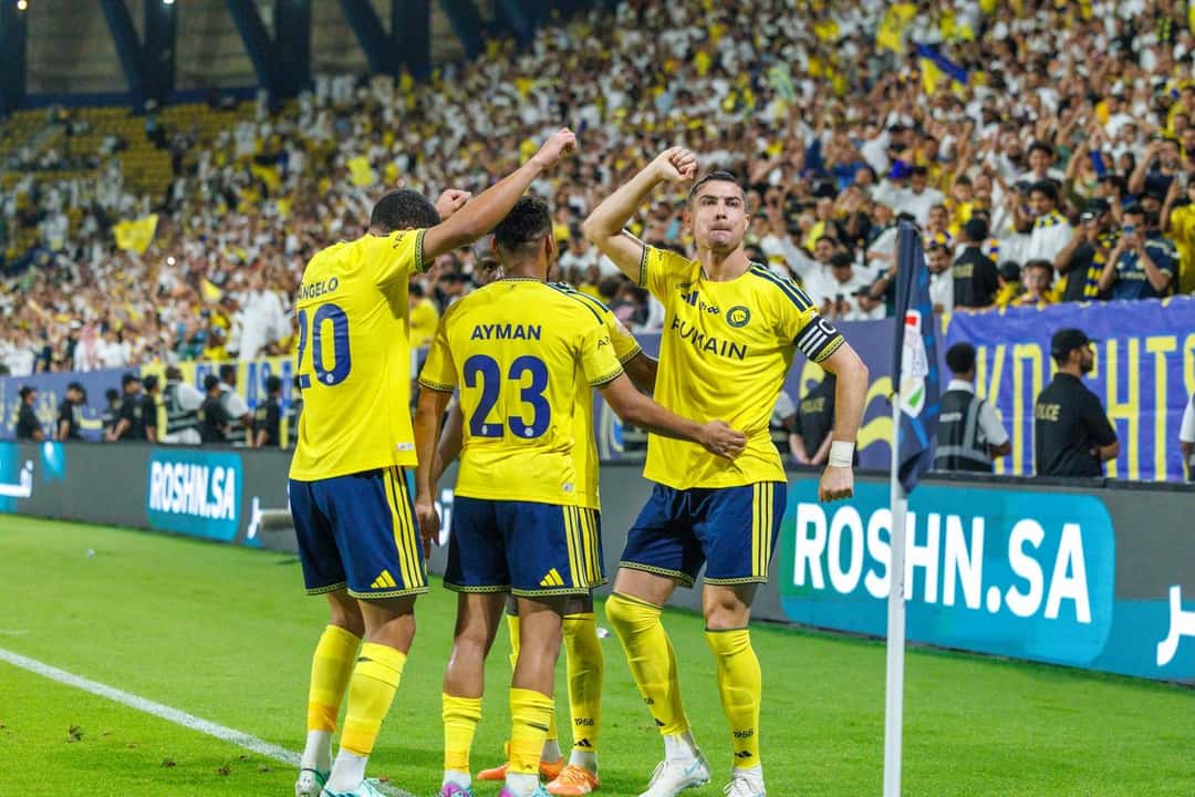 Cristiano Ronaldo, con la cinta de capitán, levanta el puño y festeja con tres compañeros del Al Nassr junto al banderín del córner, con la hinchada detrás, en el triunfo 5-3 ante el Al Kholood (foto: prensa Saudi Pro League)