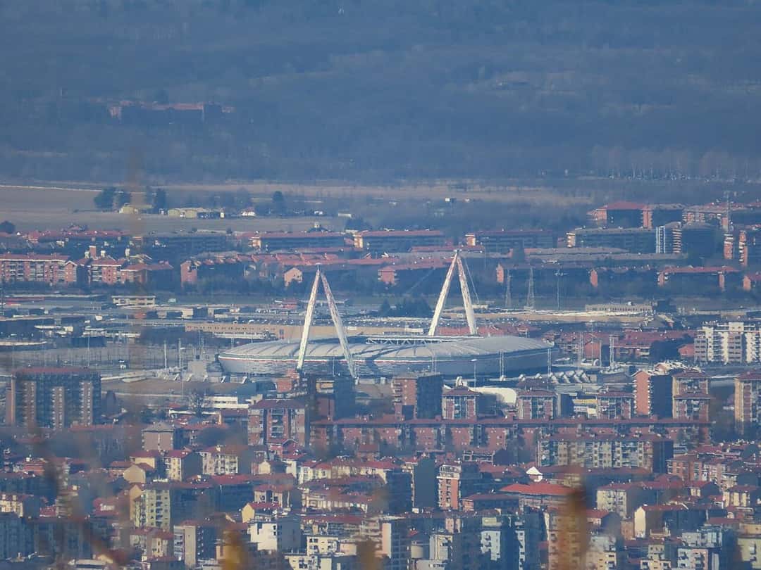 El Allianz Stadium de Turín, la cancha de la Juventus, con sus mástiles blancos, visto a la distancia entre los edificios de la ciudad; foto de archivo de marzo de 2020 (foto: Matteo Aresca 05, Wikimedia Commons, licencia CC BY-SA 4.0)