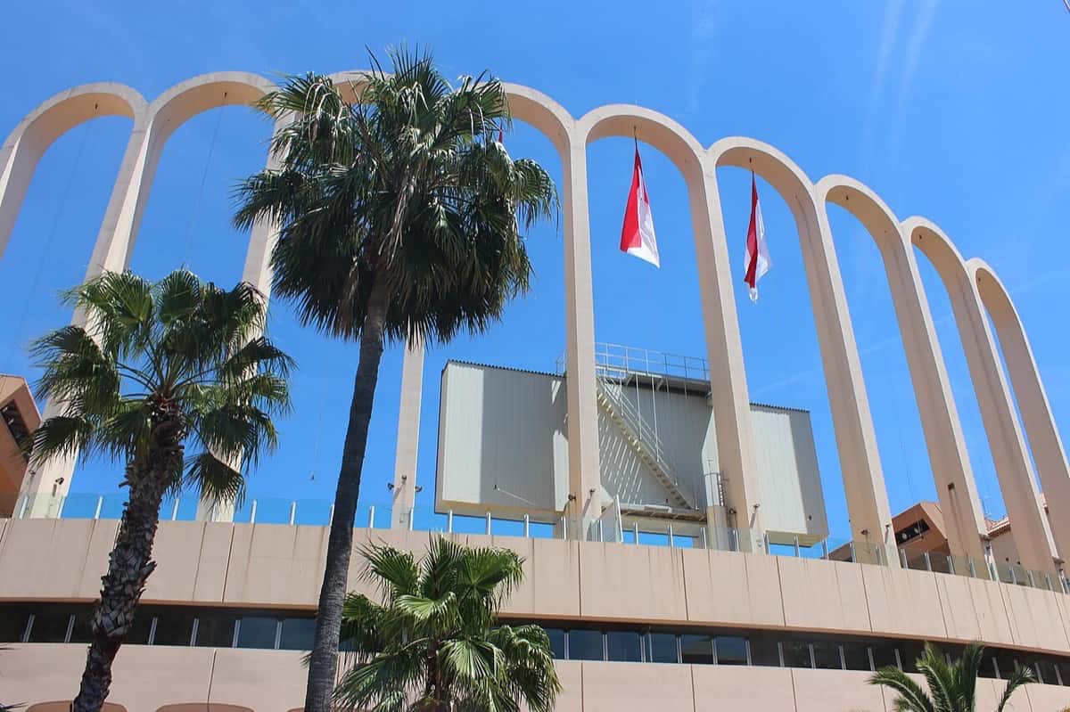 Los arcos blancos del estadio Louis II de Mónaco, con palmeras y banderas monegascas frente a la fachada; foto de archivo de mayo de 2017 (foto: Uhooep, Wikimedia Commons, licencia CC BY-SA 4.0)