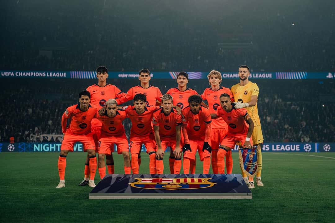 El equipo titular del Barcelona, con la camiseta naranja, posa para la foto antes del partido en el Parque de los Príncipes, con las tribunas iluminadas detrás (foto: FC Barcelona)