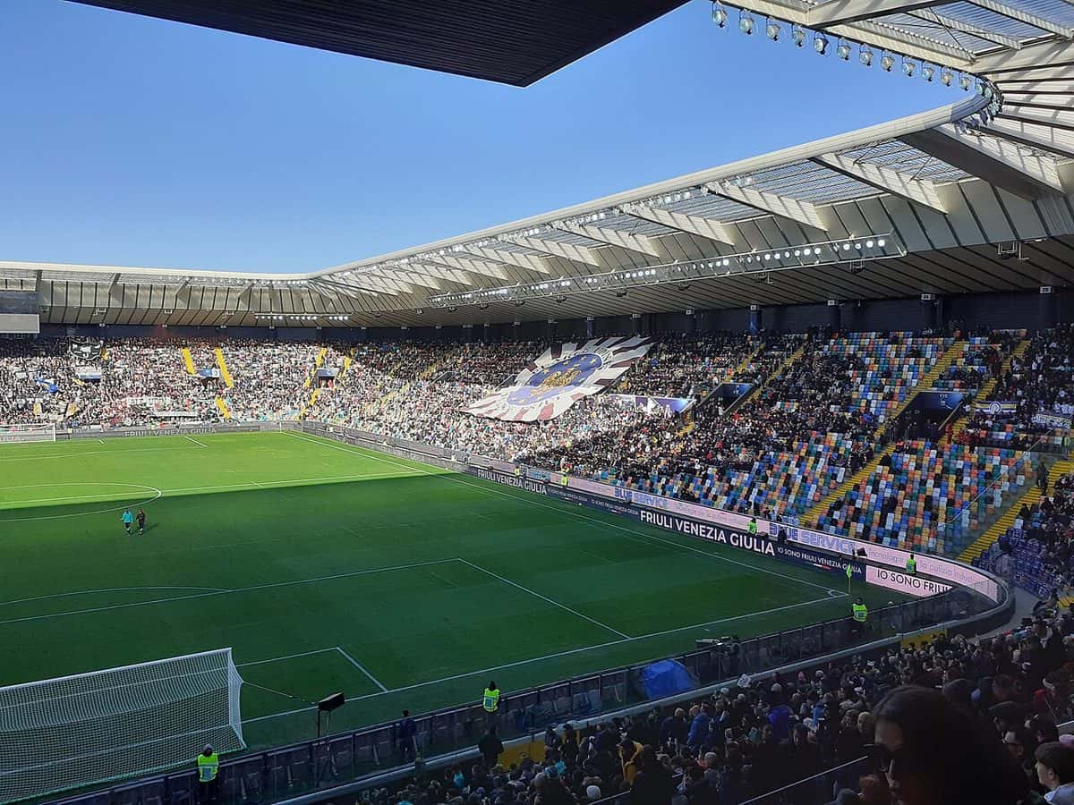 El estadio del Udinese, en Udine, con las tribunas llenas de hinchas, una bandera blanca y negra en la tribuna y la cancha a pleno sol; foto de archivo de diciembre de 2024 (foto: Holapaco77, Wikimedia Commons, licencia CC BY 4.0)