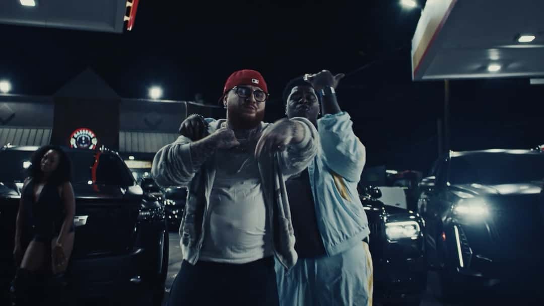 Belly Gang Kushington, de gorra roja y anteojos, y BigXthaPlug, con campera celeste, de noche en una estación de servicio con camionetas negras de fondo, en un fotograma del video oficial de «LITE»