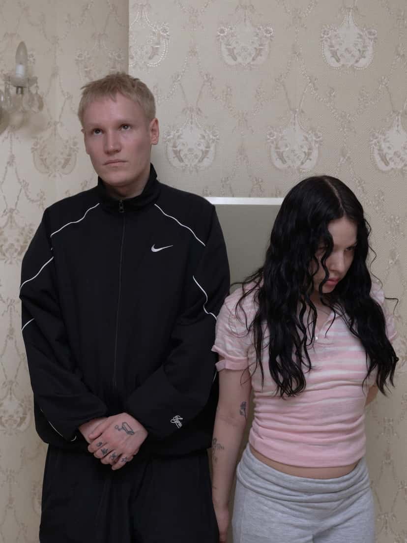 Los dos integrantes de Brutalismus 3000 posan de pie contra una pared con papel tapiz floral claro: él con campera deportiva negra y ella con remera rosa a rayas y pelo negro largo, en la foto de prensa oficial del dúo. Foto: Tom Funk