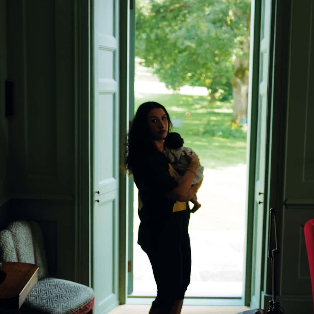 Portada oficial de «Gentlewoman» de Cleo Sol: la cantante, de pie con un bebé en brazos, junto a una puerta abierta a un jardín, en una habitación de paredes verdes