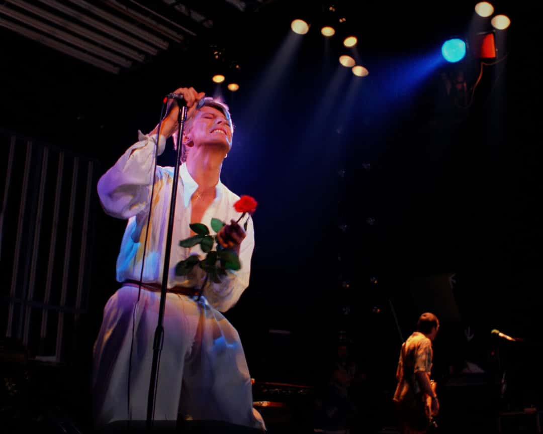 David Bowie, de camisa blanca, sostiene una rosa roja y canta con la cabeza hacia atrás junto al micrófono, en el show de 1978 (foto de prensa del proyecto «Stage»)